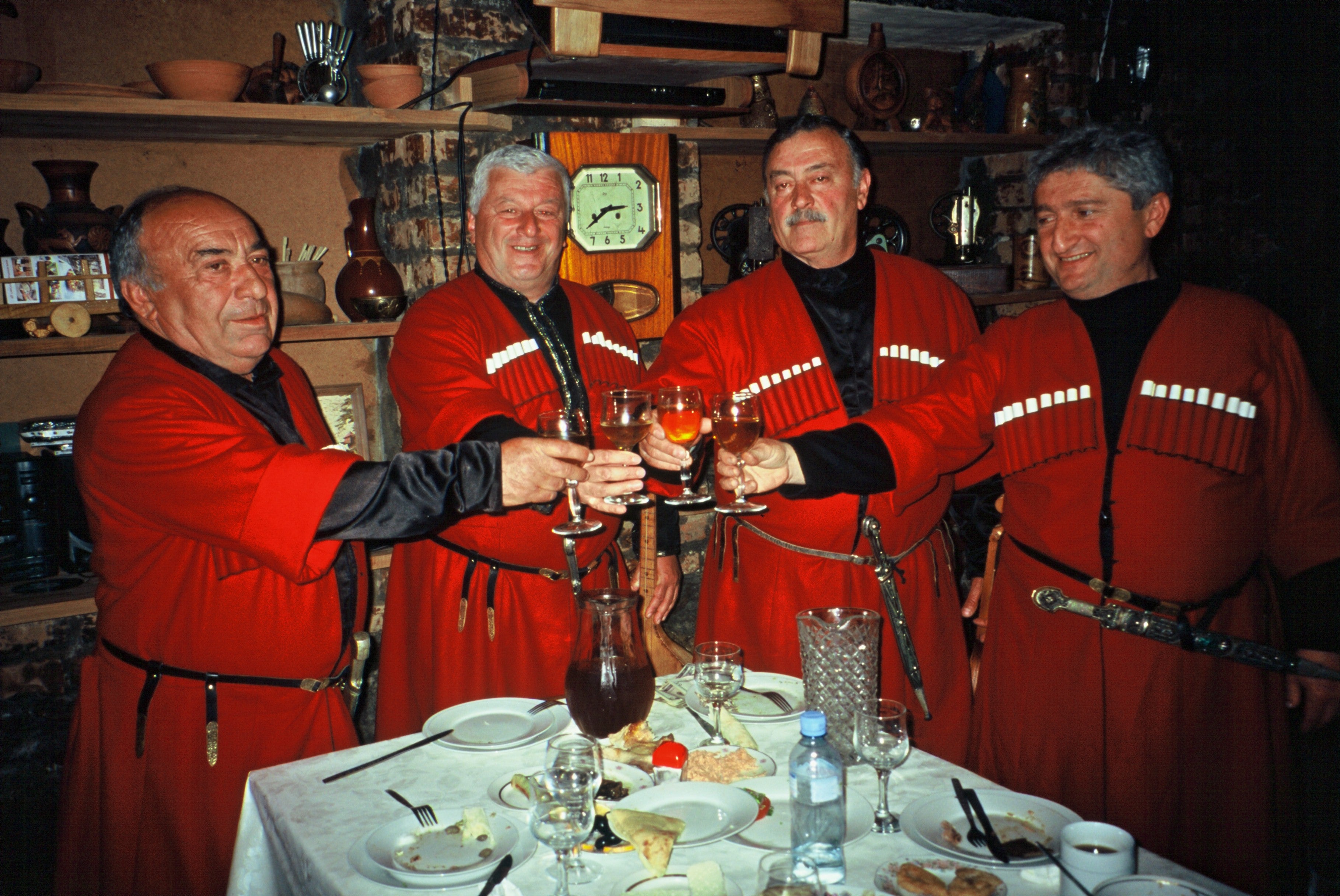 Four men in red robes toasting with glasses at a table in a dimly lit room.